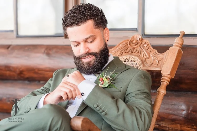 A well-dressed man with curly hair and a beard adjusts his cufflink while sitting in a rustic wooden chair. He wears a green suit with a white shirt and a small floral boutonniere.