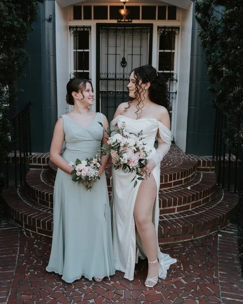 A bride in an elegant white gown and a bridesmaid in a soft green dress stand on the steps of a venue, both holding bouquets of white and blush flowers, radiating joy and elegance.