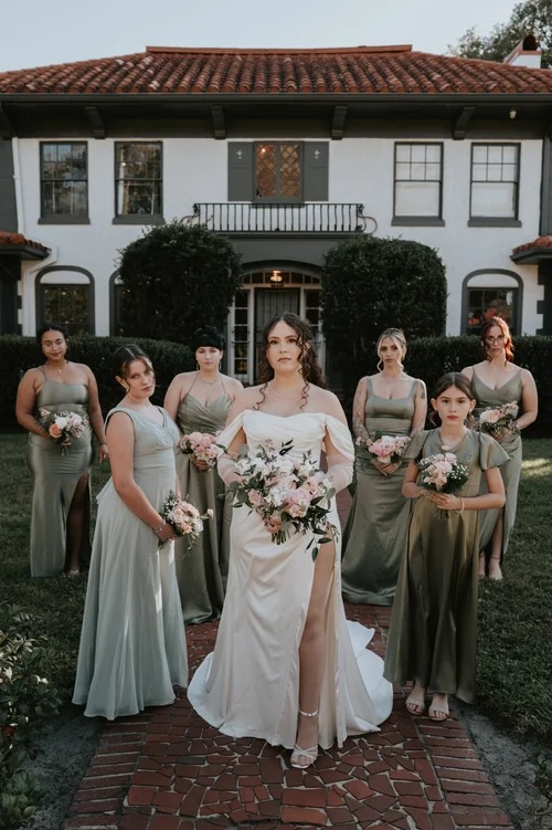 The bride in a stunning white gown stands at the front, surrounded by her bridesmaids in pastel dresses, holding bouquets of flowers outside a charming villa.