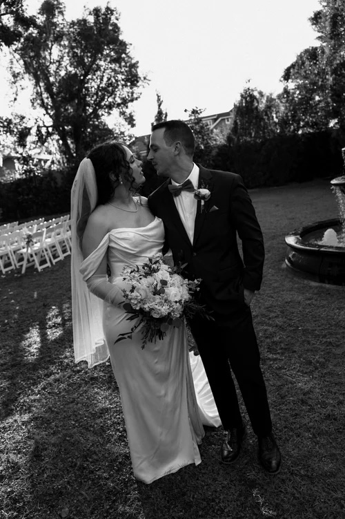 A bride in an elegant white gown, holding a bouquet of white and blush flowers, stands closely to a groom in a black suit. They share a tender moment in a garden setting, surrounded by lush greenery and a fountain in the background.