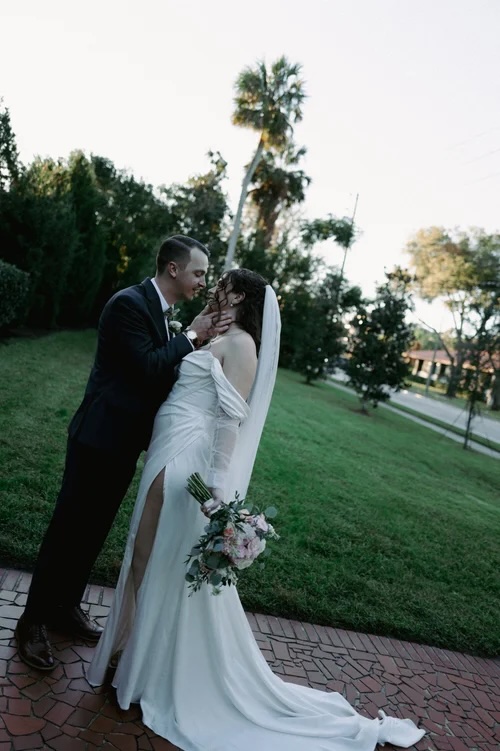 A bride in a flowing white gown and a groom in a dark suit share a romantic kiss outdoors, surrounded by lush greenery.
