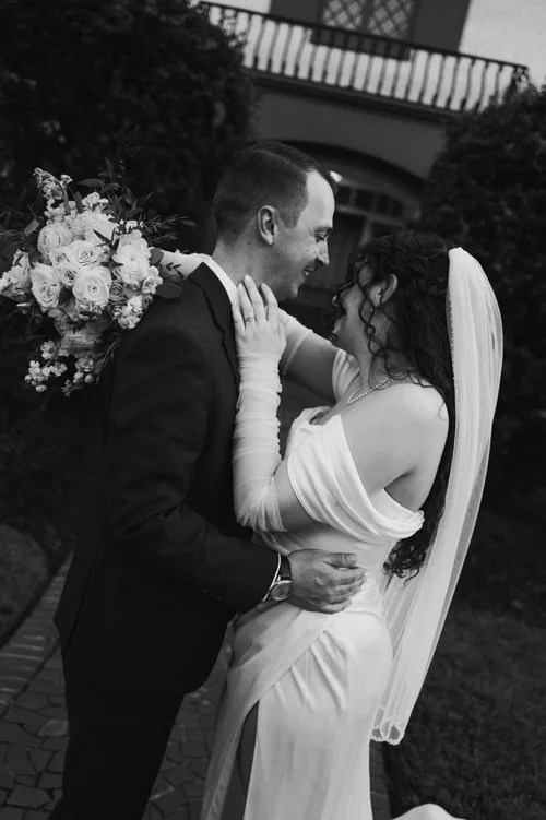 A loving couple embraces in a romantic pose, the bride wearing an elegant white gown and holding a bouquet of white and blush flowers, while the groom is dressed in a classic suit, captured in a timeless outdoor setting.