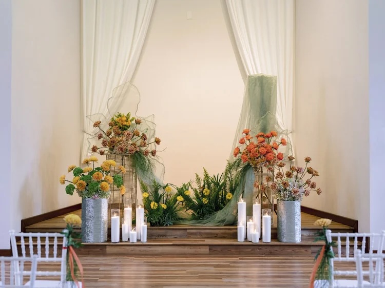 A beautifully arranged wedding altar featuring vibrant floral designs in golden yellows, oranges, and greens, complemented by candles on a wooden stage, set against a backdrop of draped fabric.