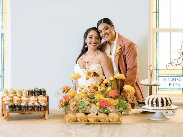 A joyful bride in a white dress stands next to a woman in a rust-colored suit, both smiling at the camera. They are surrounded by a beautifully arranged table featuring an array of desserts, including cupcakes and a cake, adorned with vibrant autumn flowers.