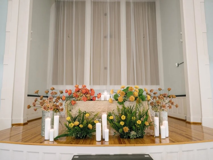 A beautifully arranged altar featuring a variety of vibrant flowers in golden yellows, oranges, and greens, along with white candles, set against a backdrop of sheer curtains.
