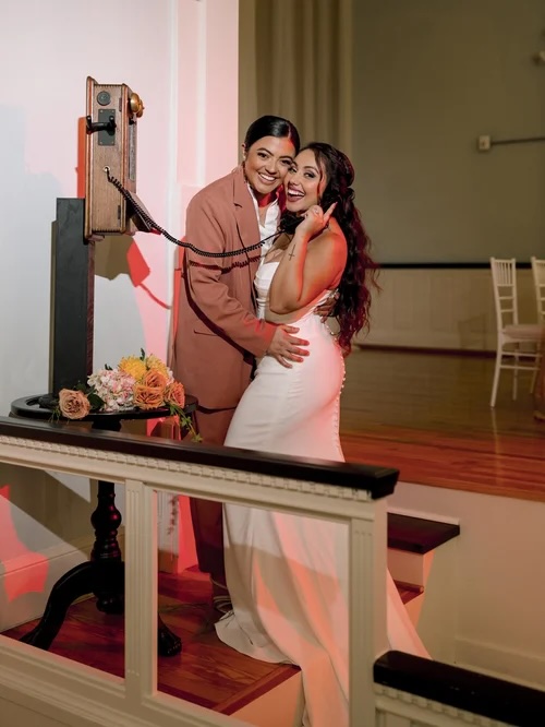 Two women joyfully posing together in a charming venue, one wearing a sleek white wedding dress and the other in a stylish blazer, with a floral bouquet on display.