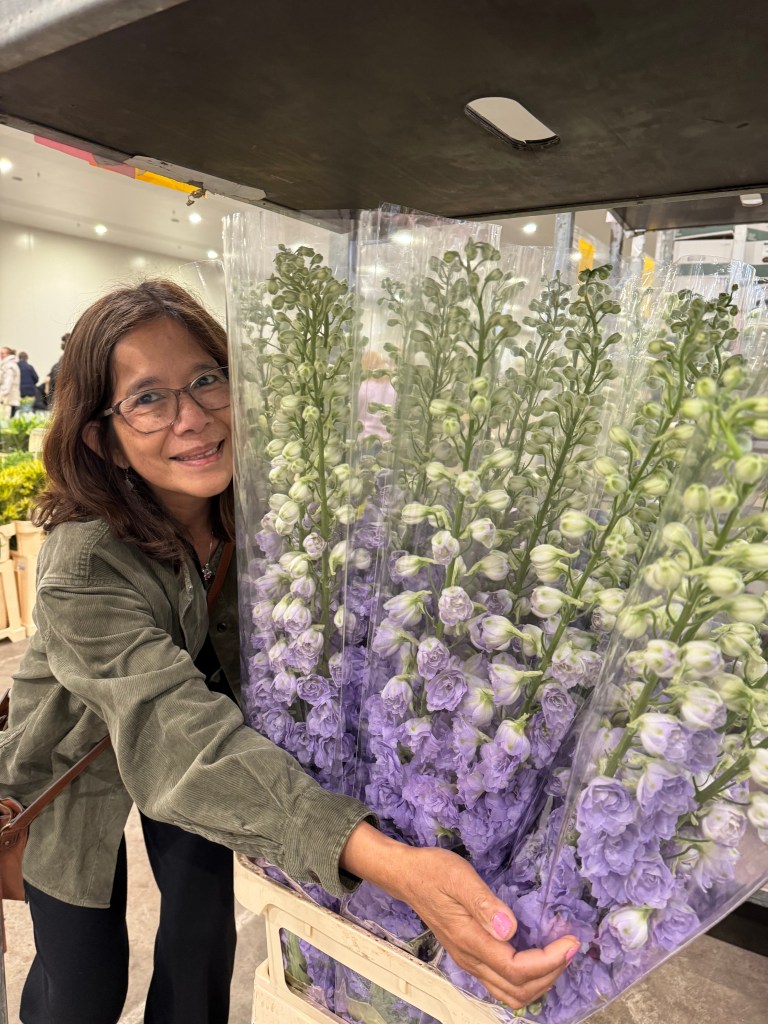 A woman with glasses smiling while holding a large bouquet of purple flowers in a flower shop.