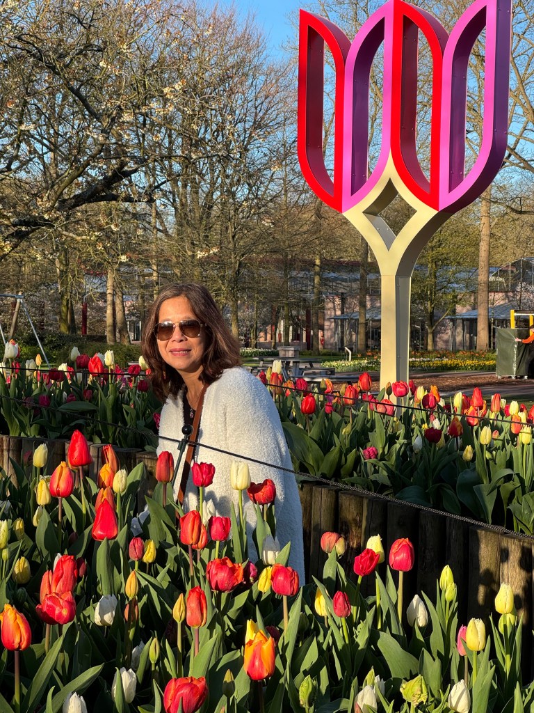 A woman with sunglasses stands among colorful tulips, with a modern pink and white sculpture in the background, under a bright blue sky.