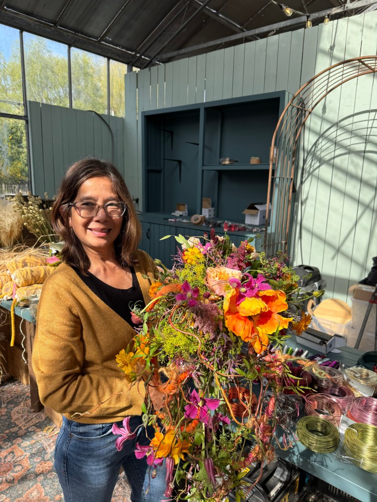 A woman stands in a bright, indoor flower shop holding a large, colorful bouquet of flowers. She is smiling and wearing a cozy sweater, with various flower arrangements and crafting supplies visible in the background.