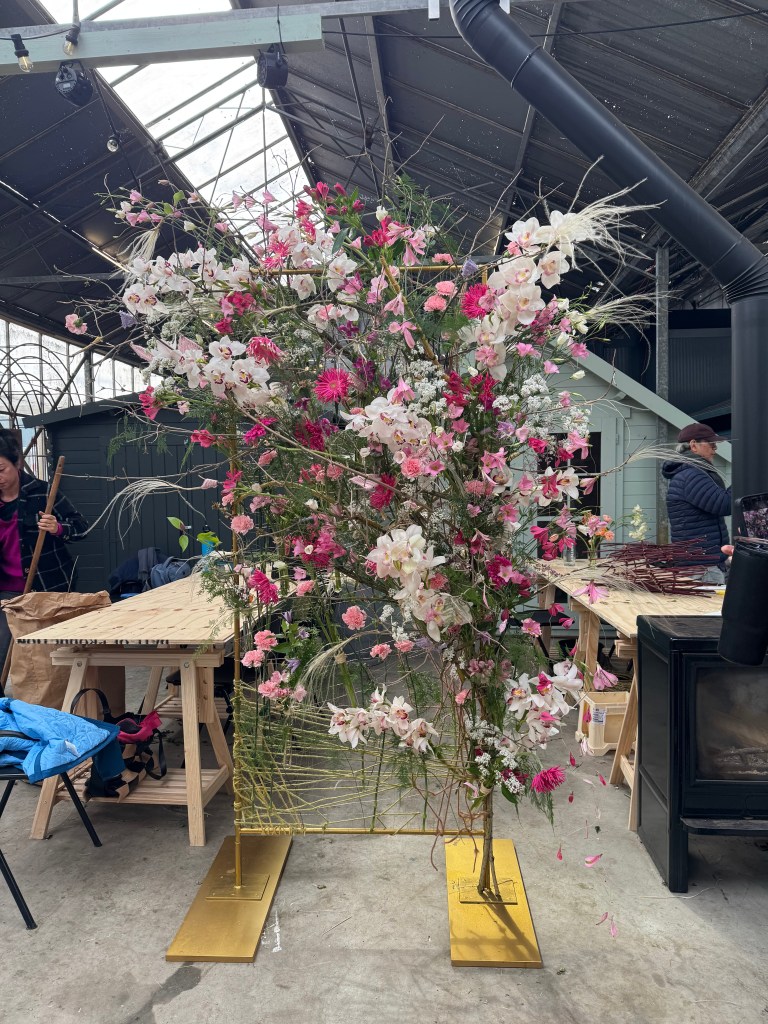 A tall floral installation featuring a variety of pink and white flowers, set against an industrial backdrop with wooden tables and people in the background.