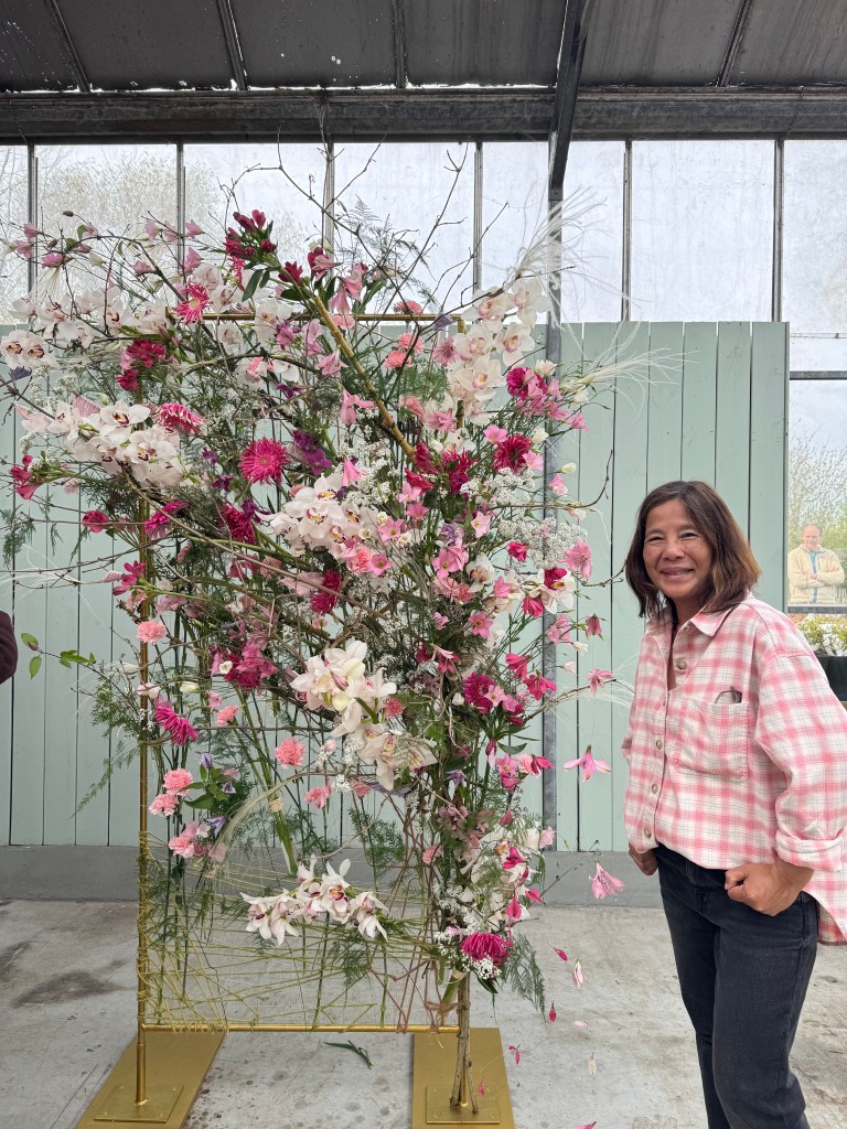 A woman smiles beside a large floral arrangement featuring pink and white flowers in a greenhouse setting.