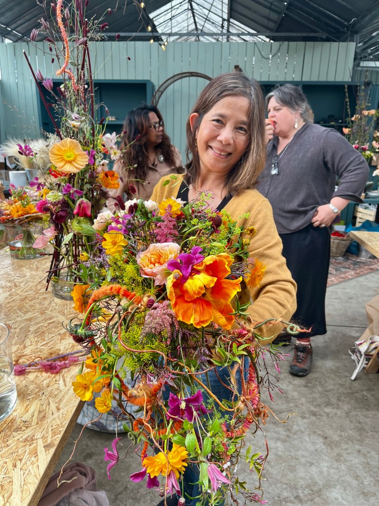 A smiling woman holding a vibrant flower arrangement filled with various colorful blooms and greenery, with a floral workshop setting in the background.